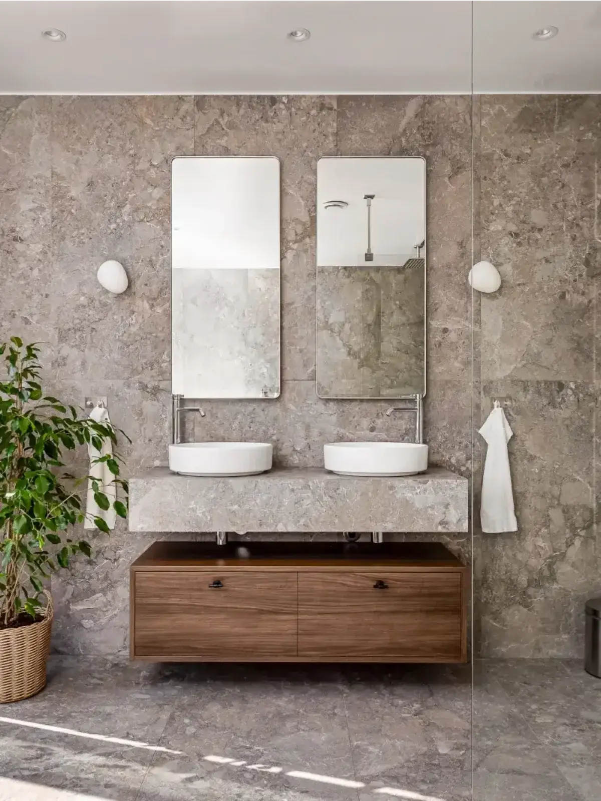 Modernt badrum med natursten i grå toner, dubbla runda tvättställ ovanpå en stenskiva, två avlånga speglar och en träkommod med lådor. En grön växt i korg ger rummet en varm och levande känsla.