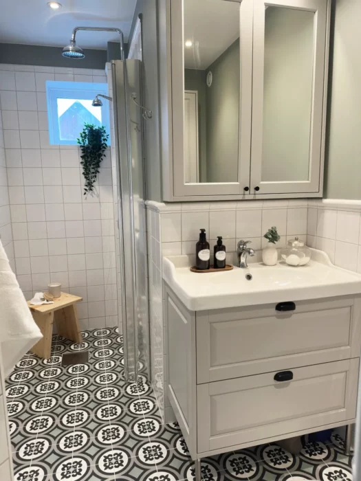 Helhetsbild av badrum i Nacka med dusch, kommod, spegel och mönstrat klinkergolv.