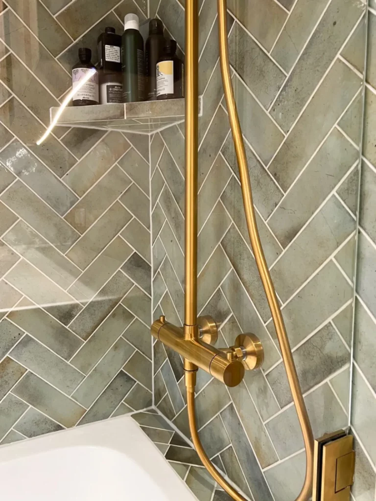 Duschhörna med mässingsdusch och grönt kakel i fiskbensmönster, hylla med flaskor i hörnet