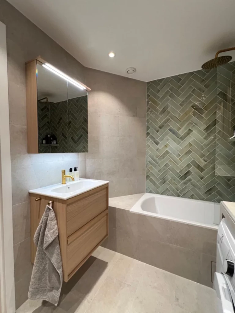 Modernt badrum med badkar, grönt fiskbenslagt kakel och träkommod med mässingsdetaljer.
