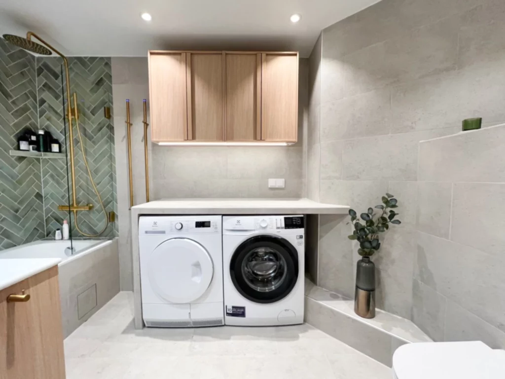 Modernt badrum med tvättmaskin och torktumlare under bänkskiva, grönt kakel i fiskbensmönster, mässingsdusch och ljusa trädetaljer