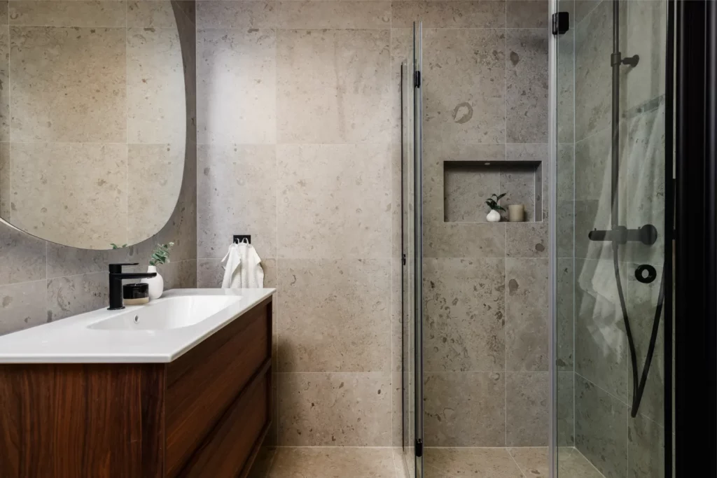 Modernt badrum efter renovering med glasdusch, stora kakelplattor i natursten, vita handdukar och stilfulla detaljer.