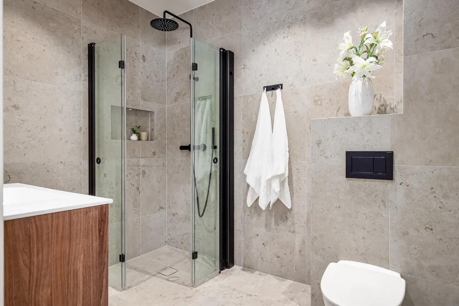 Stilrent renoverat badrum med duschhörna i glas, väggar och golv i beige sten, vägghängd toalett och inredning i trädetaljer